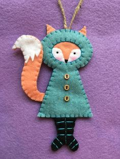 a felt fox ornament hanging on a purple background with an orange tail and black legs