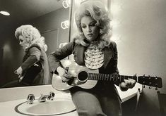 a woman sitting in front of a mirror holding a guitar next to a bathroom sink