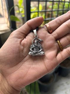 a person holding a small silver buddha charm in their left hand, with plants in the background