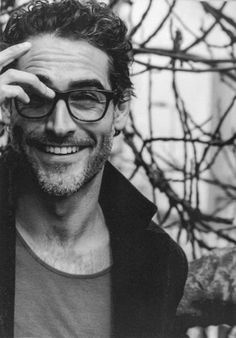 a black and white photo of a man with glasses smiling at the camera while holding his hand to his forehead