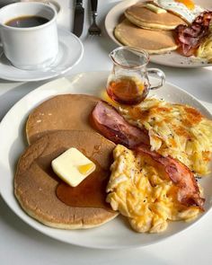 pancakes with bacon, eggs and butter are on a white plate next to two cups of coffee