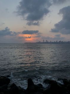 the sun is setting over the ocean with rocks in front of it and buildings in the distance