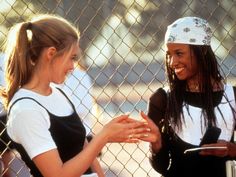 two young women standing next to each other near a chain link fence, talking and laughing