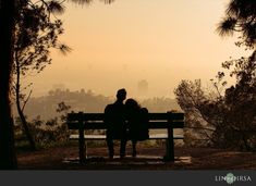 two people sitting on a bench looking at the city in the distance with trees around them