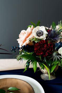 a vase filled with lots of flowers on top of a blue table cloth next to a white plate