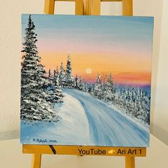 an easel with a painting on it that has a sunset in the background and snow covered trees