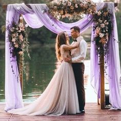 a man and woman standing under an arch with purple drapes on the water in front of them