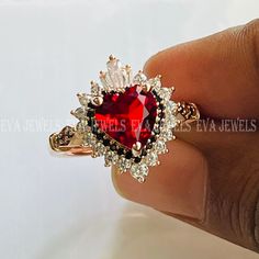 a woman's hand holding a ring with a heart shaped red stone in it