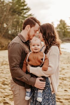 a man and woman holding a baby on the beach with trees in the back ground