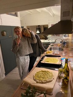 two women standing next to each other in a kitchen with pizzas on the counter