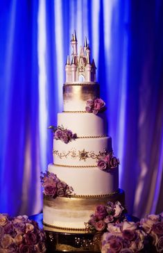 a wedding cake with purple flowers and a castle on top is sitting in front of a blue curtain