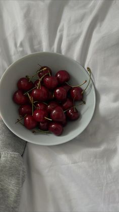 a white bowl filled with lots of cherries on top of a bed next to a person's hand