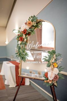 a sign with flowers and greenery on it in front of a mirror that says welcome to the wedding of tim and ash