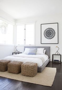 a bedroom with white walls and wood flooring has a large bed, two stools, and a painting on the wall