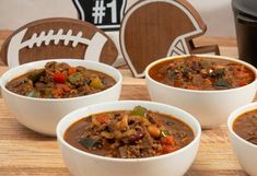 three bowls of chili and beef soup on a cutting board with a football in the background