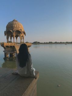 a woman sitting on the edge of a body of water looking out at a gazebo