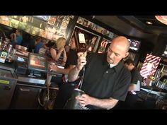 a man holding a glass in his right hand and looking at the camera while standing next to a bar