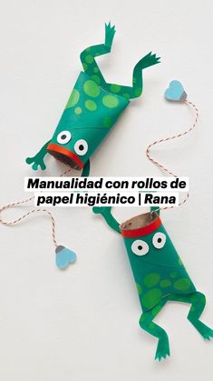 two green paper toys with eyes and legs hanging from strings on a white surface, one is shaped like a frog