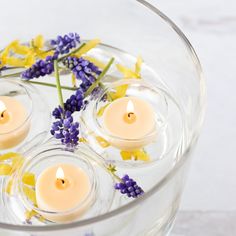 four lit candles in a glass bowl filled with water and lavenders on the side