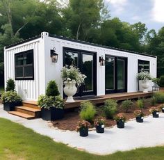 a white container home with plants and flowers on the front porch, surrounded by trees