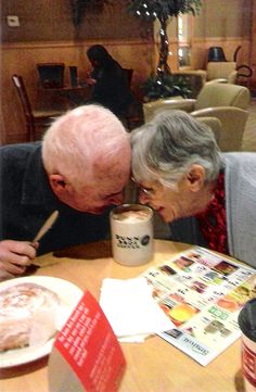 an older couple kissing each other at a table with food and drinks in front of them