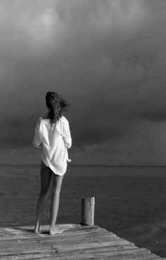 a woman standing on top of a wooden pier next to the ocean under a cloudy sky