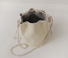 "This unique bucket bag features soft, off-white faux (fake) leather, cotton rope shoulder strap, and a drawstring closure. You can tie the cotton rope to wear this bucket bag long or short. Details: -Dimensions when laid flat: 9\" length (top to bottom), 7.5\" diameter round bottom -Shoulder strap measures approx. 45-50\" total -Soft, off-white faux (fake) leather -Solid light color interior lining -No internal pocket Because each bag is made by hand, the measurement is approximate and may vary Cream Casual Bucket Bag For Shopping, Casual Cream Bucket Bag Tote, Beige Bucket Bag With Adjustable Strap, Cream Leather Bucket Bag With Braided Handles, White Summer Bucket Bag With Dust Bag, Casual Cream Bucket Bag For Daily Use, Casual Beige Drawstring Bag For Daily Use, Casual Beige Drawstring Bag, Cream Bucket Bag With Adjustable Strap For Shopping