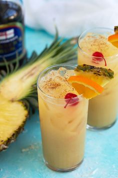 two glasses filled with fruit and garnish on top of a blue table next to a pineapple