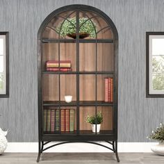 an arched glass bookcase with books and plants on it in front of two windows