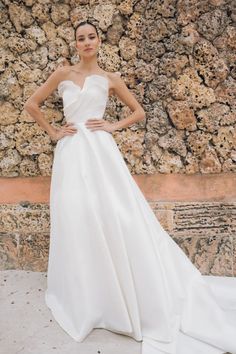 a woman standing in front of a stone wall wearing a white wedding dress with an asymmetrical neckline