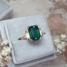 an emerald colored stone ring sitting on top of a white velvet box next to flowers