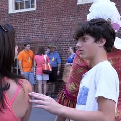 two young people talking to each other in front of a brick building while others look on