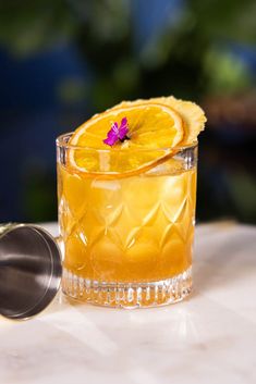 an orange drink with a purple flower on the rim sits next to a cocktail shaker