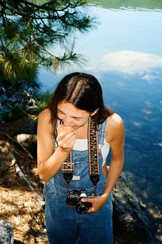 a woman standing next to a body of water holding a camera and wearing overalls