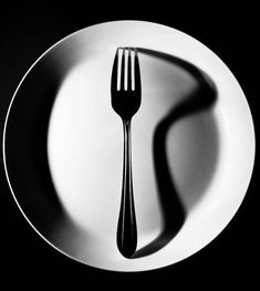 a black and white photo of a fork on a plate with the shadow of a knife