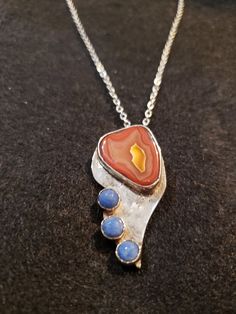 This beautiful pendant features a stunning agate stone with three Demis blue lapis on the edge of the bottom of it. The stone is set in a sterling silver bezel, creating a unique and elegant look. The pendant is handcrafted, making it a one-of-a-kind piece of jewelry. The pendant is perfect for anyone who loves unique and artisanal jewelry. The combination of the agate stone and lapis lazuli creates a beautiful and intriguing look. This pendant would make a great addition to anyone's collection, and it is sure to get compliments. Metal: Sterling Silver Gems: Agate, and 3 Demis Blue Lapis Weight: 7 grams Length: 1.75Inches Width: 0.5inches Style: Bezel Shape: Irregular Made in Ohio, USA Artisan Agate Cabochon Necklace, Amber Sterling Silver Necklace With Cabochon, Collectible Agate Pendant Jewelry, Artisan Lapis Lazuli Pendant Necklace, Silver Pear-shaped Cabochon Jewelry, Ohio Usa, Blue Lapis, On The Edge, Agate Stone