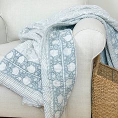 a white couch with a blue and white blanket on it's back next to a wicker basket