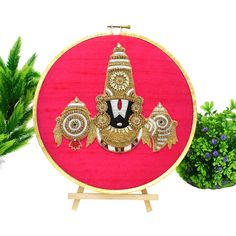 a red and gold embroidered art work on a wooden stand next to potted plants