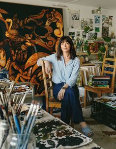 a woman sitting on a chair in front of a table with many paintings and brushes