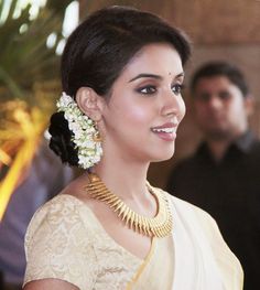 a woman in a white sari and gold necklace with flowers on her head is smiling at the camera