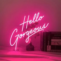 a pink neon sign that says hello gorgeous next to two books on a table with a plant in the corner