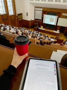 a person holding a red coffee cup in front of an auditorium full of people sitting at desks