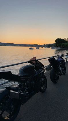 two motorcycles parked next to each other on the side of a road near water with boats in the background