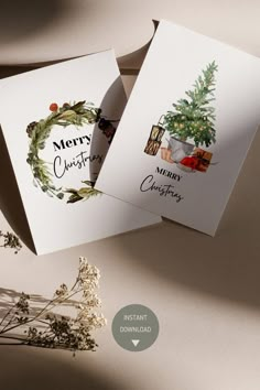 two christmas cards sitting next to each other on top of a table with flowers and greenery