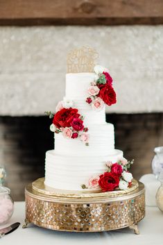 a white wedding cake with red and pink flowers on the top is sitting on a gold stand