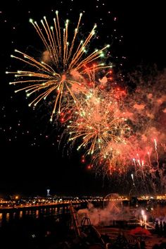 fireworks are lit up in the night sky over water and buildings with lights on them