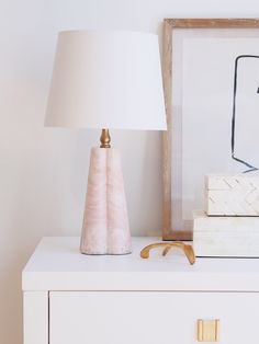 a pink lamp sitting on top of a white dresser next to a framed photograph and a pair of glasses