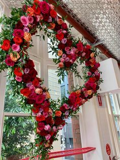 a heart shaped wreath with red and pink flowers hanging from it's sides in front of a window