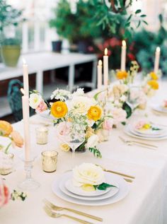 the table is set with white and yellow plates, silverware, flowers and candles