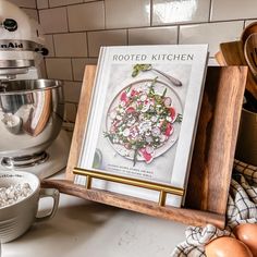 a cookbook sitting on top of a wooden tray next to eggs and an mixer